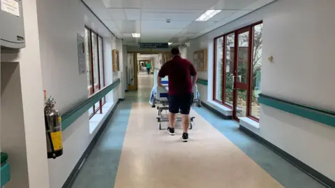 A man in a red t-shirt and shorts pushing a hospital bed through a hospital corridor, walking away. The walls are white, with blue hand rails through them, and large windows along the corridor.