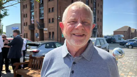 A close up shot of Vic, wearing a grey polo shirt. He has grey hair and is smilling into the camera.