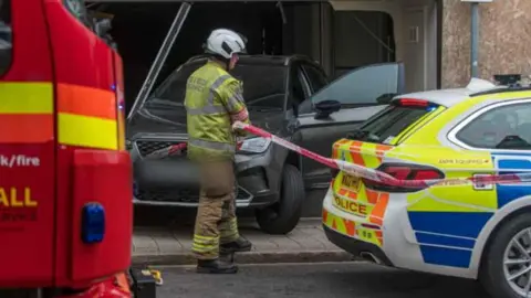 Emergency services person in front of a car and holding some tape and a police car.