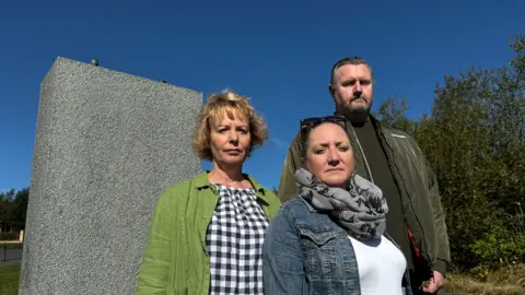 A woman wearing a green top stands next a woman wearing a blue shirt, and a man wearing a green jacket, all solemn-faced. Behind them is a grey column where the stolen figure was displayed.