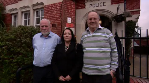 Maxine Pooley is pictured in a dark coloured coat, stood alongside two male volunteers outside Pinner police station. The man on her right has a beard and a blue shirt and the man on her left has a grey, green and blue striped top