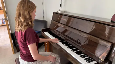 Freya is sitting at a glossy brown piano, with her left hand resting on the keys. Her right hand is resting on her knee, which is where it normally remains while she is performing. She is sitting upright on the piano stool and is wearing a burgundy t-shirt and white trousers with thin blue stripes and a pink dotted pattern. 