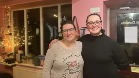 Polly Harris and Lisa Myatt standing in the Communitea hub in Wigton. In the background is a lit-up Christmas tree and the shops window with items to be sold as part of their thrift shop.