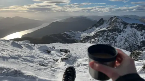 Aishling Burke A hand holds a cup overlooking snow-covered peaks and a bright sky.