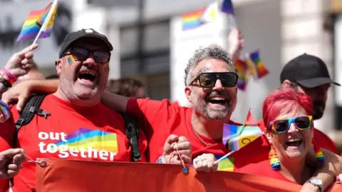 PA Media A group of people waving rainbow LGBTQ+ flags and wearing rainbow accessories hold a large red banner.
