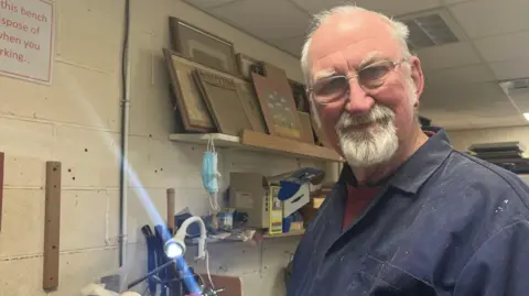 A man standing beside a woodturning lathe in a workshop, looking at the camera. The person is wearing glasses and a dark work shirt with small wood shavings on it. The lathe and a small adjustable lamp are visible in the foreground. Behind the person are shelves holding tools and boxes. 