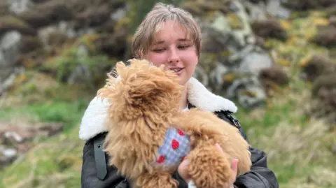 A woman with tied-back hair, wearing a brown leather jacket, is holding a light brown coloured dog in her arms.