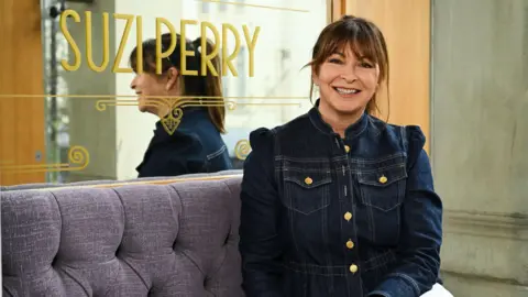 Broadcaster and Journalist Suzi Perry sits on a purple sofa in a theatre in front of a mirror which has her name printed on it. She wears a denim jacket and smiles at the camera.
