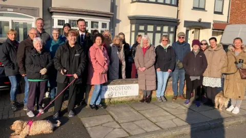 Twenty-one people are standing on the street, Kingston Avenue, and are looking at the camera. They are wearing coats of varying colour and some are squinting from the sun. They are positioned around the street name sign on a brick wall, in front of two semi-detached houses.