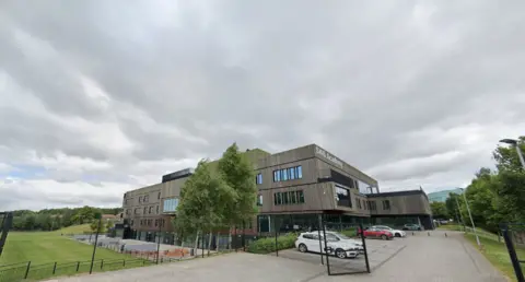 School which has light brown walls and had playing fields on one side. There are also trees and several cars outside. 