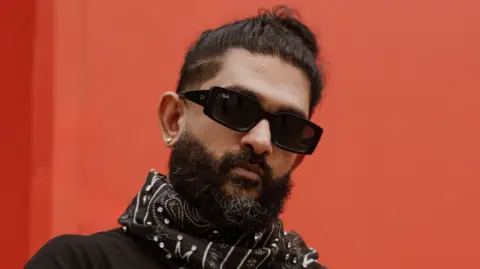 Soozana Pvan Sid Sriram, a male singer wearing black sunglasses, standing in front of a red box.