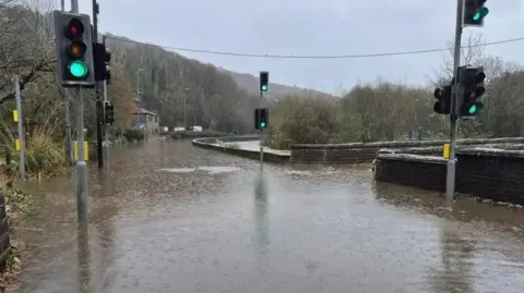 West Yorkshire Police Burnley Road at Callis Bridge is shown heavily flooded as water covers the entire road. Traffic lights are shown as is a stone bridge which crosses the River Calder.