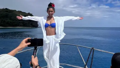 JENNIFER CHAN Seorang wanita mengenakan kemeja dan bawahan putih serta atasan bikini biru berpose di atas perahu di laut, di depan seorang pria yang tengah mengambil gambar dengan ponselnya.