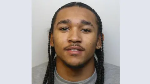 A police mugshot of Joel Binnings. He is looking at the camera wearing a grey sweatshirt and has long dreadlocks