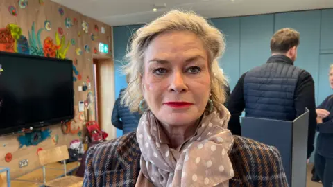 Blonde-haired woman looking at the camera. She is wearing a scarf and is wearing a tweed like jacket.