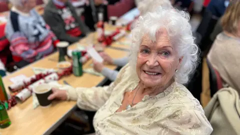 Jackie Holden has curly white hair and is wearing a sparkly gold top. She is sitting at a table, holding her drink and smiling at the camera. 