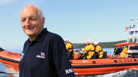 RNLI/ Ian Foster David John Haines standing to the left wearing a navy blue T-shirt with a orange lifeboat and crews on board. He is smiling at the camera and the T-shirt has a RNLI badge on the right. The lifeboat crew are all wearing yellow suits. 