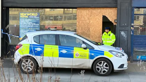 A female police officer wears a hi-vis jacket and black hat as she guards a vacant shop, which has been partially boarded up. She stands behind a hatchback police car and next to blue and white tape.