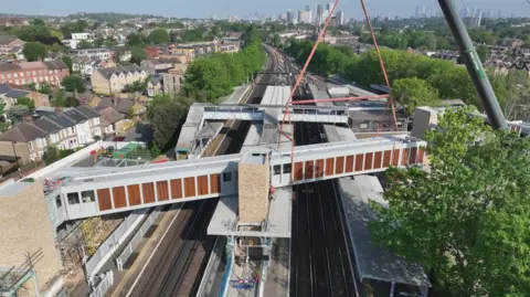 The footbridge attached to a crane being lifted and placed on top of Hither Green station. 