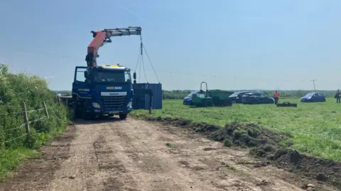 A lorry with a crane carrying a shipping container. It is on a dirt path within a field. A group of cars are on the grass alongside two people in the distance.