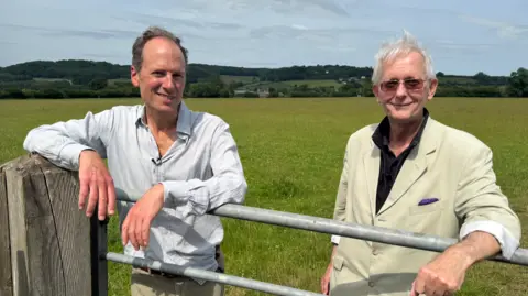 The two men are leaning on a gate. In the background are the farm's meadows, with hills beyond. 