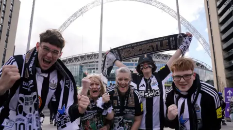 Five Newcastle United fans on Wembley Way ahead of the Carabao Cup final at Wembley Stadium, London. They are cheering and waving scarves.