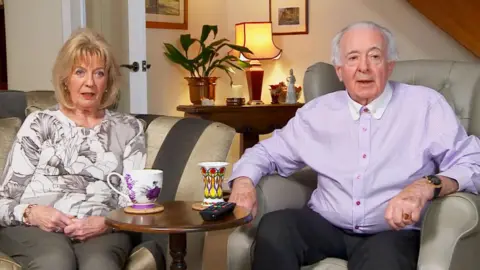 Ken Harwood and his wife Anne sitting on armchairs in their living room in an episode of Gogglebox. Ken Harwoos has short white hair and is wearing a pale pink shirt and dark trousers, a watch and gold ring. Anne Harwood has blonde hair in a bob and blue eyes. She is wearing a white and grey top with a floral pattern and grey trousers. The pair are watching a TV, which is out of shot. In front of them there is a coffee table with a remote control and two colourful China cups.