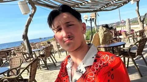 Gloucestershire Police Tyler Durn is wearing a red and white shirt and is sat by the sea at a restaurant table looking into the camera. He has short dark hair, short facial hair and an earring in one ear.