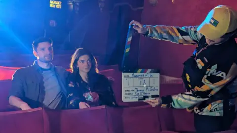 Callum McCourt, TMM Productions A man and a woman sit on red cinema chairs. A man wearing a yellow hat and patterned jacket holds a clapboard in front of them.