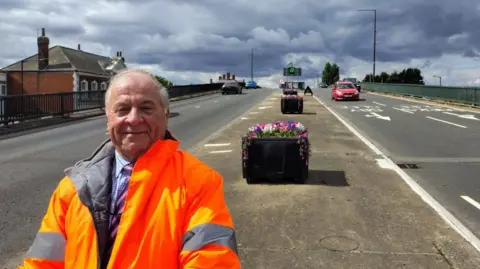 North East Lincolnshire Council A man with grey hair wears a suit under an orange hi-vis jacket and stands in front of a planter in the central reservation on a dual carriageway. There are several black planters behind him featuring colourful artificial flowers. 