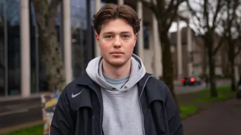 A brown-haired boy staring straight into the camera, he stands in front of a modern glass building and dotted trees. He wears a black jacket with a grey jumper underneath.
