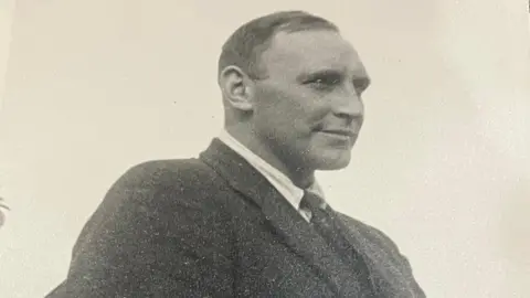 A black and white image of Blair 'Paddy' Mayne. He is looking away from the camera and is wearing a black blazer, white shirt and tie. 