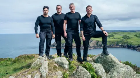PA Media Four men in dark shirts stood on cliff edge with the ocean in the background