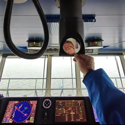 YouTube Un hombre con gafas se mira en un pequeño espejo en un equipo en el puente de mando de un gran ferry.