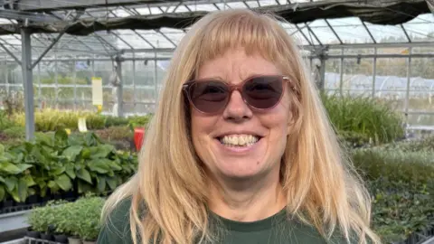A smiling Heather Lewis standing in the plant nursery with tables full of plants in the background. She has long blond hair and dark glasses.