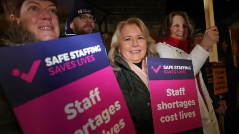 PA Media Pat Cullen, a woman with shoulder-length blonde hair, smiles as she joins a picket outside a hospital in the dark in Newcastle, England. There are two women stood either side of her holding purple and pink signs saying 'Safe staffing saves lives. Staff shortages cost lives' and one man with glasses standing behind her.