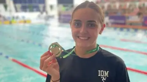 LEE HOLLAND Laura Kinley wears a black T-shirt, which has the letters IOM and a triskelion in white to her left. She holds a gold medal attached to a green ribbon that is draped around her neck. There is a swimming pool behind her.