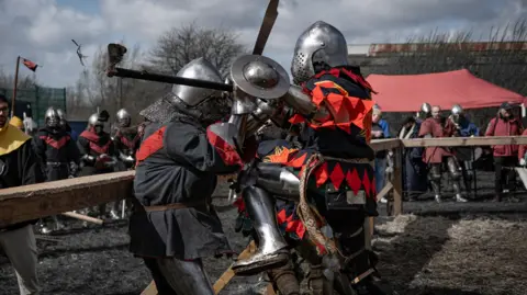 Two people wearing medieval suits of armour engaged in combat, with a crowd of people watching behind