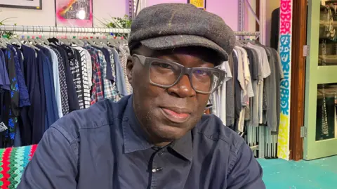 Kwame Daniels is wearing a grey hat and blue shirt. He has black glasses. There is a rail of clothing behind him and colourful posters on the wall.