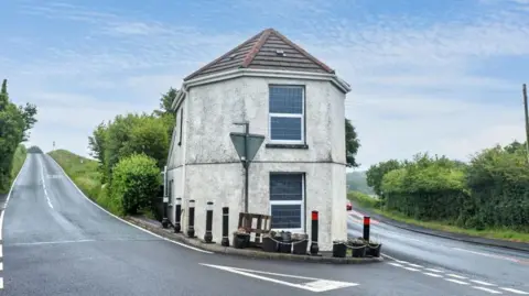 A small white building with large windows is situated at the corner of a road. It was a former toll building and has two main roads either side. 