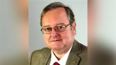 A man with short, greying hair, wearing black framed glasses, a green suit jacket with a white shirt and red tie, looks towards the camera with a neutral expression on his face