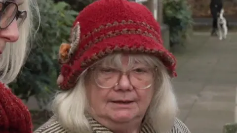 Mary Pimm in a red knitted hat. She wears glasses and a striped coat.