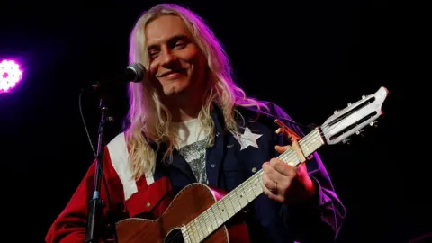 Casey Lowery, who has long blonde hair, holding a brown wooden guitar on stage