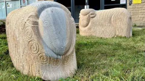 Two large carved stone sheep stand on a patch of grass in front of a brick building, each decorated with swirling patterns cut into their bodies. One sheep is darker and more detailed, while the other is lighter and simpler, with signs and windows from nearby offices visible behind them.