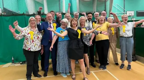 A group of about 20 people, men and women, celebrating winning an election. They are of all different ages and all are wearing yellow rosettes and cheering with their arms up in a sports hall. 