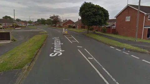 A Google street view of a residential road - there is a grass verge on the left and a row of red brick houses on the right.