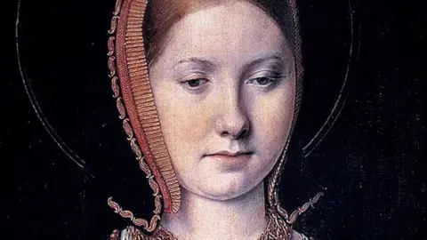 Getty A close-up of a portrait of Katharine of Aragon as a young woman. She is wearing a black headdress edged with red and gold braid and embroidery. Her red hair can be seen pulled back at her forehead. She is lowering her eyes to look down. 
