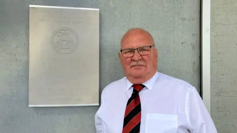 BBC/Olivia Richwald A man in a shirt and tie stands in front of the plaque which has the names of the six soldiers on it. There is also an image of the regimental badge and writing in Flemish. 