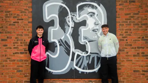 Millie Evans Two young men with dark hair standing in front of a brick wall beside a black and white sign with a large number 33 on it. One of the young men has a pink running top and the other a white running top