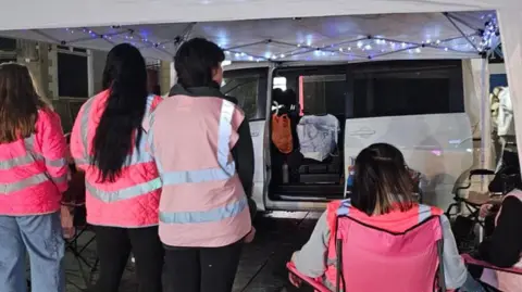 Women's Street Watch The volunteers photographed from behind, underneath their white marquees, which has been assembled next to their white van.
